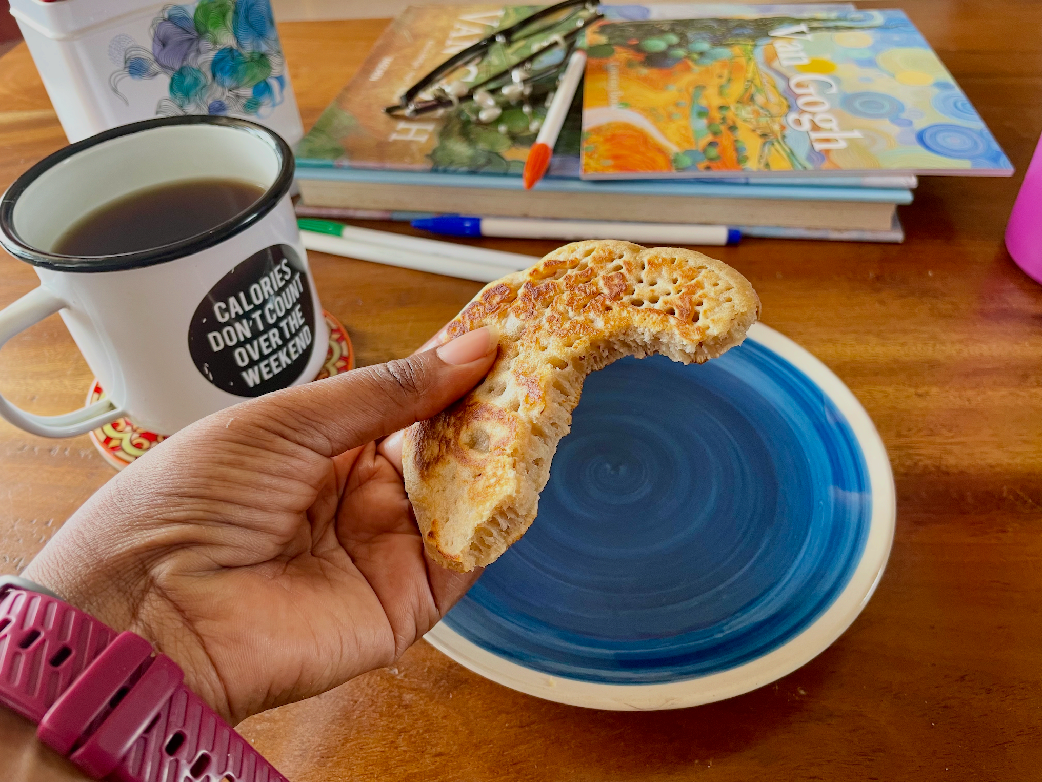 A hand holds a partially eaten homemade pancake above a blue plate, showing its golden-brown surface and fluffy interior. A mug of tea, colouring books, and art supplies sit on a wooden table in the background.