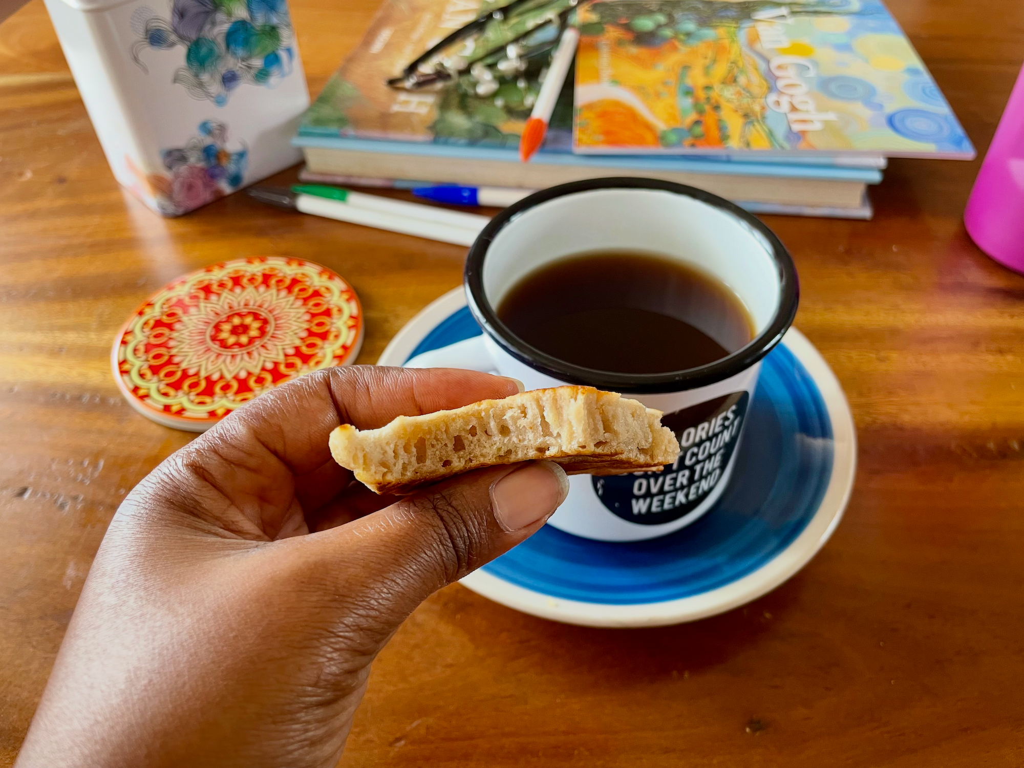 A hand holds a piece of homemade pancake, showing its soft, airy interior. A mug of black tea sits on a blue saucer in the background, alongside colouring books, pens, and art supplies on a wooden table.